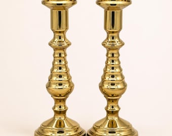 May include: Two gold-colored candle holders. Each holder has a wide, round base, a long, textured stem, and a small, circular top to hold a candle. The candle holders are made of a shiny, metallic material.