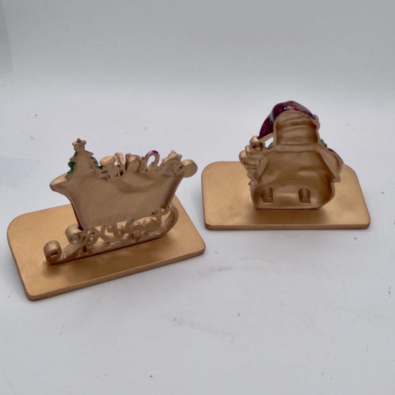 May include: Two gold-colored metal decorative figures on rectangular bases. One figure depicts a sleigh with a small tree, while the other shows the back view of a figure wearing a hat with a dark red finish. Both items have a smooth, matte gold finish.