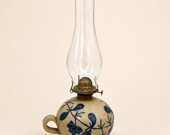 Könnte beinhalten: Eine Öllampe aus Keramik mit einem runden beigen Sockel, der mit handgemalten blauen Blumenmustern und einem kleinen seitlichen Griff versehen ist. Die Lampe besitzt einen messingfarbenen Metallbrenner und einen hohen, klaren Glaszylinder.