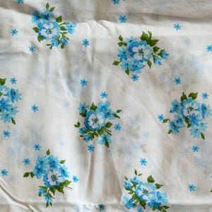 May include: White fabric with a repeating pattern of blue floral bouquets and small blue flowers. The bouquets have a central cluster of white flowers surrounded by blue petals and green leaves. The fabric is slightly wrinkled.