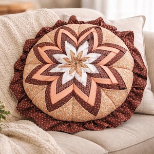 May include: A round, quilted pillow with a floral design. The pillow features a central starburst pattern in white, peach, and brown tones, surrounded by a ruffled edge. The pillow is placed on a beige sofa.