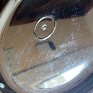 Op de afbeelding: Een cirkelvormig, gepolijst zilverkleurig metalen deksel met een centrale verhoogde ronde knop. Het reflecterende oppervlak vertoont zichtbare fijne krassen en lichte slijtage over de gehele koepel, met een gladde, gebogen rand.