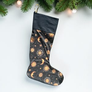 May include: Black Christmas stocking with a celestial pattern of suns, moons, stars, and compass roses in gold. The stocking has a black cuff and a gold loop for hanging. Festive holiday decor.