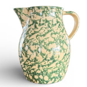 May include: A ceramic pitcher with a cream-colored base and a green, speckled pattern. The pitcher has a spout for pouring and a curved handle. The design is reminiscent of vintage kitchenware.