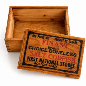 May include: An open, rectangular wooden box with a lid. The lid displays text in red and black, including "FINAST BRAND BONELESS SALT CODFISH" and "FIRST NATIONAL STORES, BOSTON, MASS." The box is made of light-colored wood.