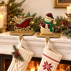 May include: Two gold-toned metal stocking holders featuring a red sleigh filled with toys and a snowman wearing a red hat and green scarf. Each holder supports a cream-colored knit stocking, one with a pine and berry accent and one with a red snowflake.