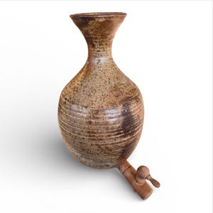Puede incluir: Jarra de agua de cerámica marrón hecha a mano, con una forma bulbosa única y un cuello estrecho. La jarra tiene un grifo y un asa de madera para dispensar. La superficie tiene una textura moteada y líneas horizontales.