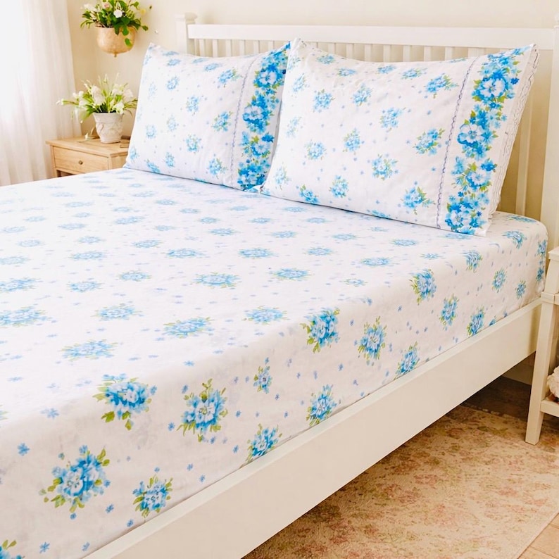 May include: White bed linens with a blue floral pattern. The set includes a fitted sheet and two pillowcases. The floral design features clusters of blue flowers with green leaves. The bed frame is white.