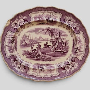 May include: An oval ceramic platter with a scalloped edge, featuring a purple transferware print on a cream background. The design depicts a landscape with figures on horseback, a tent, a deer, and architectural structures, framed by floral borders.