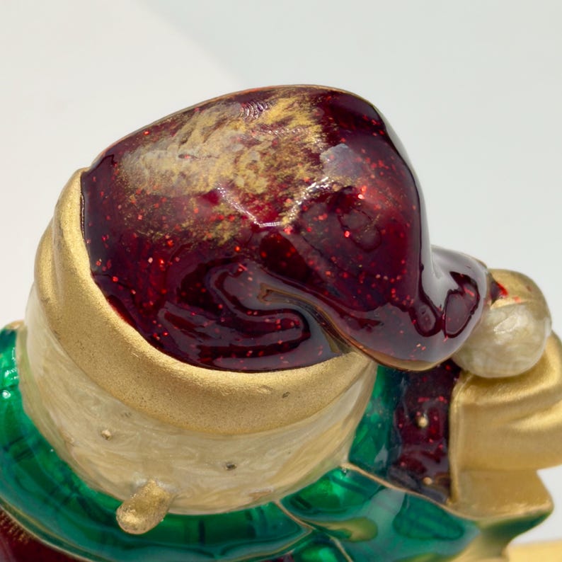 May include: A decorative figurine featuring a deep red, glittery hat with a gold band and a white pom-pom. The figure has a cream-colored textured head and a green collar. The surface of the hat has a glossy, enamel-like finish with gold accents.
