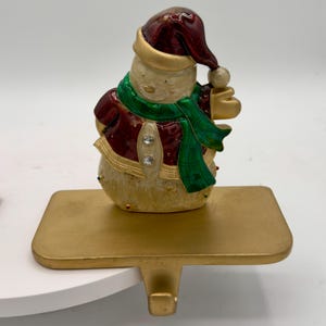 May include: A decorative stocking holder featuring a snowman figurine wearing a deep red hat and jacket with a green plaid scarf. The snowman has a cream-colored body with gold accents and two clear crystal buttons. It sits on a gold metal base.