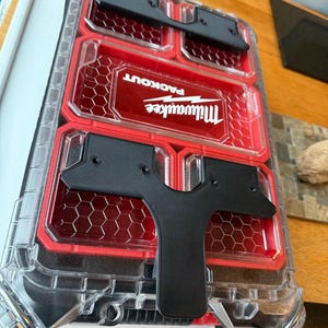 May include: A clear and red Milwaukee Packout tool organiser with black handles. The organiser has multiple compartments and a honeycomb pattern design. The Milwaukee logo and Packout text are visible on the front.