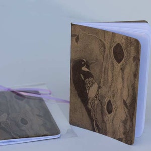 May include: Two brown notebooks with a wood grain pattern and a black and white woodpecker illustration. The notebooks are wrapped in clear plastic with a purple ribbon.