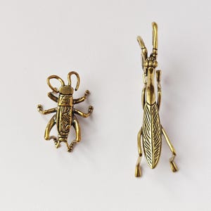 Puede incluir: Dos figuras de insectos de latón. Una es un pequeño grillo detallado con patas y antenas extendidas. La otra es una langosta estilizada más grande con un cuerpo texturizado y patas largas, de pie.