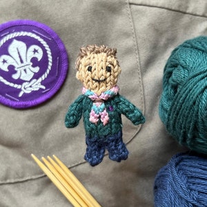 May include: A knitted figure of a person wearing a green and blue scout uniform with a purple scout badge and a pink scarf. The figure is sitting on a brown fabric surface with two balls of yarn, one green and one blue.