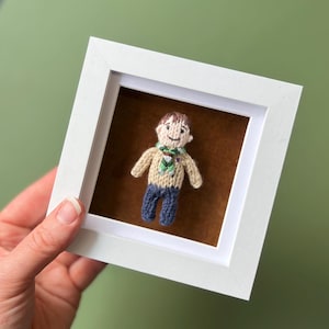 May include: A knitted figurine of a person wearing a brown shirt, green scarf, and blue trousers. The figurine is displayed in a white frame with a brown mount.