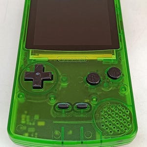 Puede incluir: Una consola de juegos port&aacute;til verde transparente con pantalla negra, cruceta y botones. El texto "RETROPIXEL" es visible. La consola tiene un altavoz y una toma de auriculares.