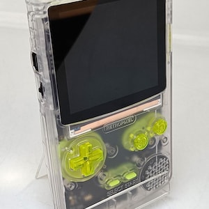 Puede incluir: Un dispositivo de juego port&aacute;til transparente con botones y detalles en verde ne&oacute;n. El dispositivo tiene una pantalla negra y el texto "RETROPIXEL" es visible. El dispositivo est&aacute; sobre un soporte transparente.
