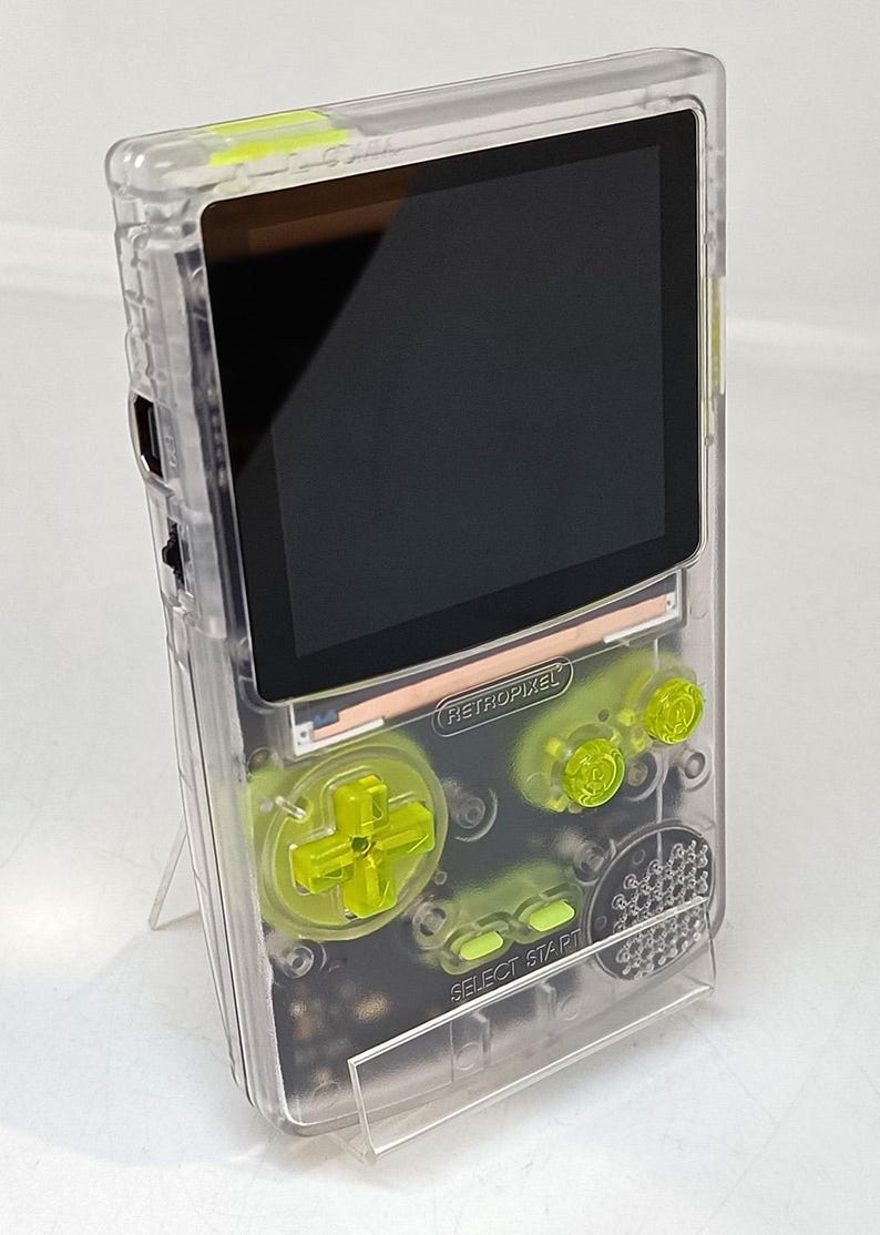 Puede incluir: Un dispositivo de juego port&aacute;til transparente con botones y detalles en verde ne&oacute;n. El dispositivo tiene una pantalla negra y el texto "RETROPIXEL" es visible. El dispositivo est&aacute; sobre un soporte transparente.