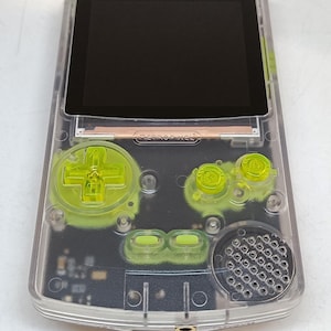 Puede incluir: Una consola de juegos port&aacute;til transparente con una pantalla negra y botones verde ne&oacute;n. El dispositivo tiene un pad direccional, botones y un altavoz. El texto "retropixel" es visible.
