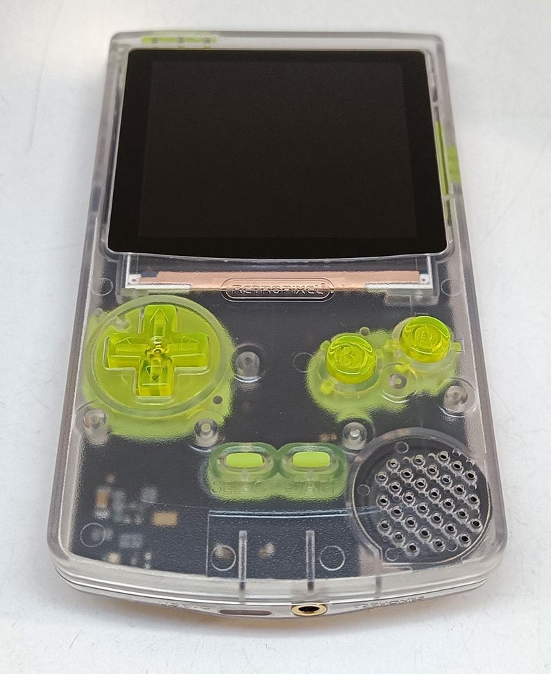 Puede incluir: Una consola de juegos port&aacute;til transparente con una pantalla negra y botones verde ne&oacute;n. El dispositivo tiene un pad direccional, botones y un altavoz. El texto "retropixel" es visible.