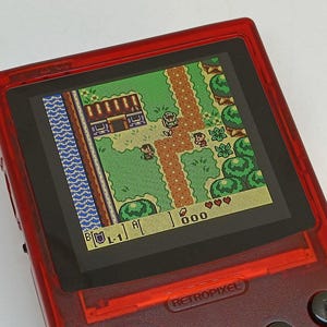 Puede incluir: Una consola de juegos port&aacute;til roja que muestra una escena de videojuego retro. La pantalla muestra una vista desde arriba de un mundo pixelado con personajes, edificios y un camino. La consola tiene botones y el texto "RETROPIXEL".