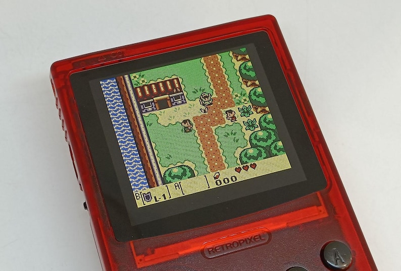 Puede incluir: Una consola de juegos port&aacute;til roja que muestra una escena de videojuego retro. La pantalla muestra una vista desde arriba de un mundo pixelado con personajes, edificios y un camino. La consola tiene botones y el texto "RETROPIXEL".