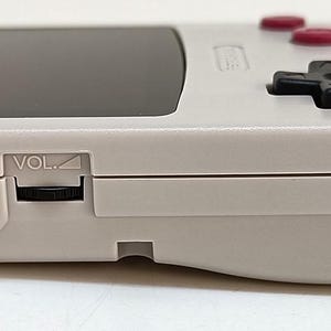May include: A light gray handheld gaming console with a black screen, red buttons, and a black directional pad. The side view shows ports labeled "EXT" and "VOL".