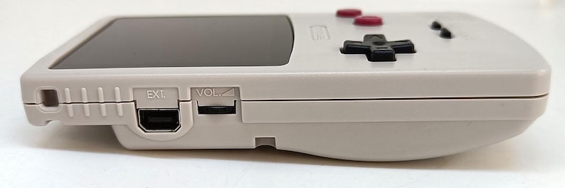 May include: A light gray handheld gaming console with a black screen, red buttons, and a black directional pad. The side view shows ports labeled "EXT" and "VOL".
