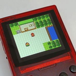 Puede incluir: Una consola de juegos port&aacute;til roja que muestra una escena de videojuego retro. La pantalla muestra personajes pixelados en un paisaje con un edificio, &aacute;rboles y agua. El dispositivo tiene botones y el texto "RETROPIXEL".