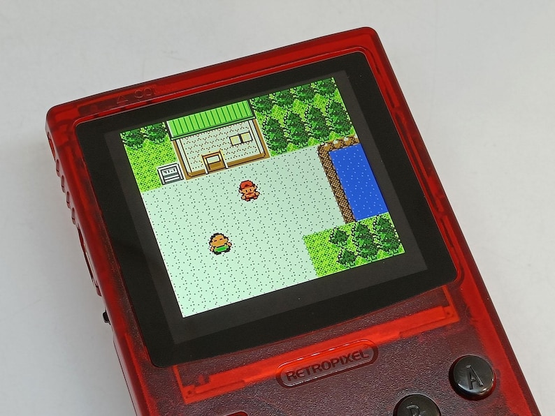 Puede incluir: Una consola de juegos port&aacute;til roja que muestra una escena de videojuego retro. La pantalla muestra personajes pixelados en un paisaje con un edificio, &aacute;rboles y agua. El dispositivo tiene botones y el texto "RETROPIXEL".