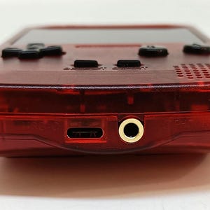 May include: A close-up of a transparent red handheld gaming console. The device has black buttons, a screen, and a headphone jack. The bottom edge features a USB-C port and a gold-colored headphone jack.