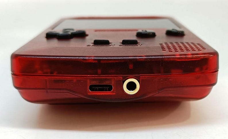 May include: A close-up of a transparent red handheld gaming console. The device has black buttons, a screen, and a headphone jack. The bottom edge features a USB-C port and a gold-colored headphone jack.