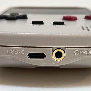 May include: A handheld gaming console in a light gray color. The bottom edge features a USB-C port, a headphone jack, and the word "PHONES" printed next to the jack. The console has black directional buttons and red action buttons.
