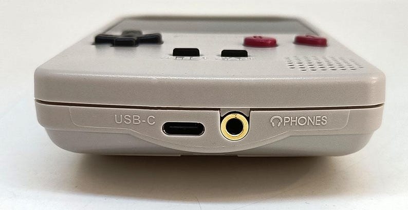 May include: A handheld gaming console in a light gray color. The bottom edge features a USB-C port, a headphone jack, and the word "PHONES" printed next to the jack. The console has black directional buttons and red action buttons.