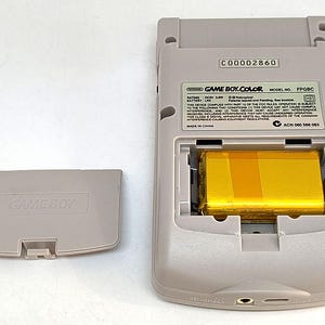 May include: A light gray Game Boy Color handheld game console with the battery compartment open, revealing a gold-colored battery. The battery cover is detached and lying to the left. The console has the text "GAME BOY.COLOR" and "FPGBC".