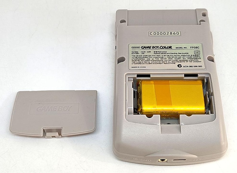 May include: A light gray Game Boy Color handheld game console with the battery compartment open, revealing a gold-colored battery. The battery cover is detached and lying to the left. The console has the text "GAME BOY.COLOR" and "FPGBC".