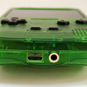 Puede incluir: Una consola de juegos port&aacute;til verde transparente. El dispositivo presenta una pantalla negra, un mando direccional negro y botones negros. El borde inferior tiene un puerto USB-C, una toma de auriculares y la palabra "PHONES".