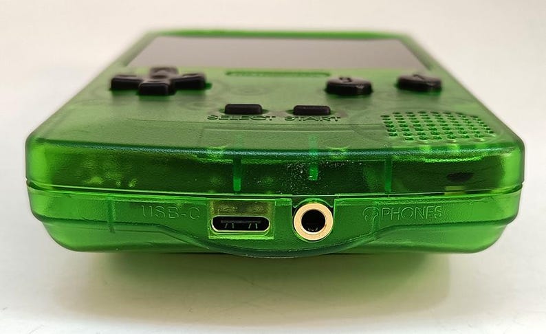 Puede incluir: Una consola de juegos port&aacute;til verde transparente. El dispositivo presenta una pantalla negra, un mando direccional negro y botones negros. El borde inferior tiene un puerto USB-C, una toma de auriculares y la palabra "PHONES".