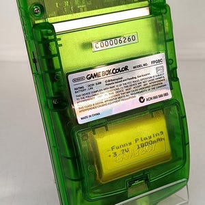 Puede incluir: Una consola de juegos port&aacute;til Game Boy Color verde transparente. La parte trasera del dispositivo es visible, mostrando los componentes internos y un compartimento de bater&iacute;a. El texto "GAME BOY.COLOR" y "Funny Playing" son visibles.