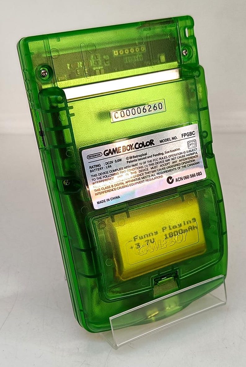 Puede incluir: Una consola de juegos port&aacute;til Game Boy Color verde transparente. La parte trasera del dispositivo es visible, mostrando los componentes internos y un compartimento de bater&iacute;a. El texto "GAME BOY.COLOR" y "Funny Playing" son visibles.