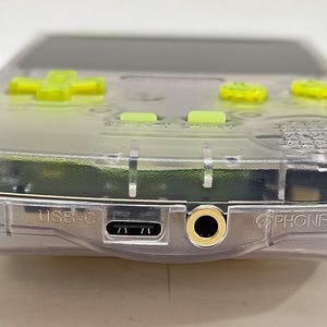 Puede incluir: Una consola de juegos port&aacute;til transparente con botones verde ne&oacute;n y un mando direccional. El dispositivo tiene un puerto USB-C y una toma de auriculares. La palabra "PHONES" est&aacute; impresa en el lateral.