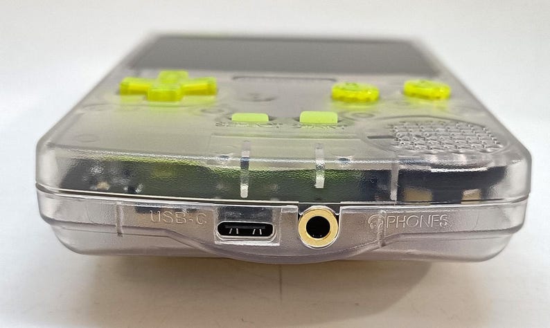 Puede incluir: Una consola de juegos port&aacute;til transparente con botones verde ne&oacute;n y un mando direccional. El dispositivo tiene un puerto USB-C y una toma de auriculares. La palabra "PHONES" est&aacute; impresa en el lateral.