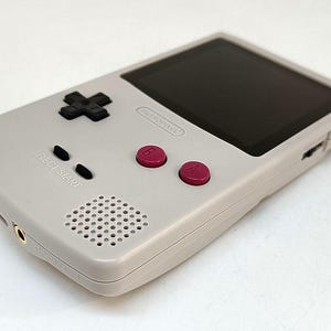 May include: A handheld gaming console in a light gray color. It features a black directional pad, black start and select buttons, and two magenta buttons labeled A and B. The screen is black, and the console has a speaker and a headphone jack.