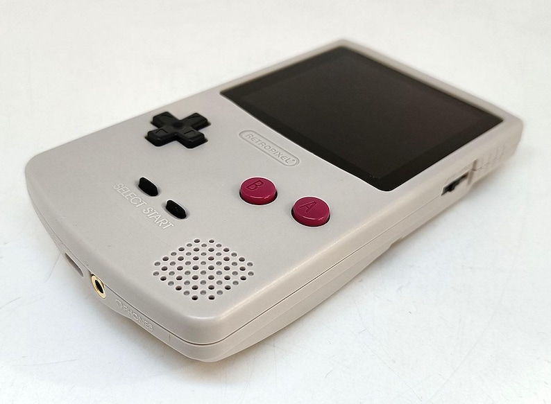 May include: A handheld gaming console in a light gray color. It features a black directional pad, black start and select buttons, and two magenta buttons labeled A and B. The screen is black, and the console has a speaker and a headphone jack.