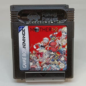 Puede incluir: Un cartucho de Game Boy Advance para el juego "Mother 1+2". El cartucho es negro con una etiqueta roja que presenta personajes de dibujos animados. Las palabras "Mother 1+2" se muestran de forma destacada. El cartucho también tiene el texto "Game Boy Advance" y "Funny Playing."