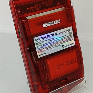 May include: A transparent red Game Boy Color handheld game console. The back of the console has a sticker with the text "GAME BOY COLOR" and other technical details. The console is on a clear plastic stand.