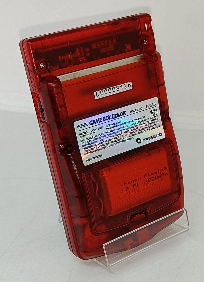 May include: A transparent red Game Boy Color handheld game console. The back of the console has a sticker with the text "GAME BOY COLOR" and other technical details. The console is on a clear plastic stand.