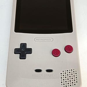 May include: A light gray handheld gaming console with a black screen and red A and B buttons. The console has a directional pad, select and start buttons, and a speaker. The text "RETROPIXEL" is printed on the console.