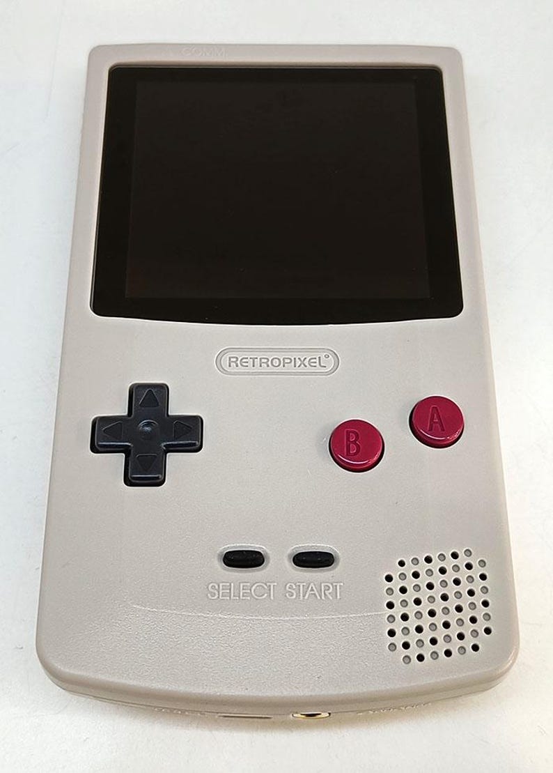 May include: A light gray handheld gaming console with a black screen and red A and B buttons. The console has a directional pad, select and start buttons, and a speaker. The text "RETROPIXEL" is printed on the console.